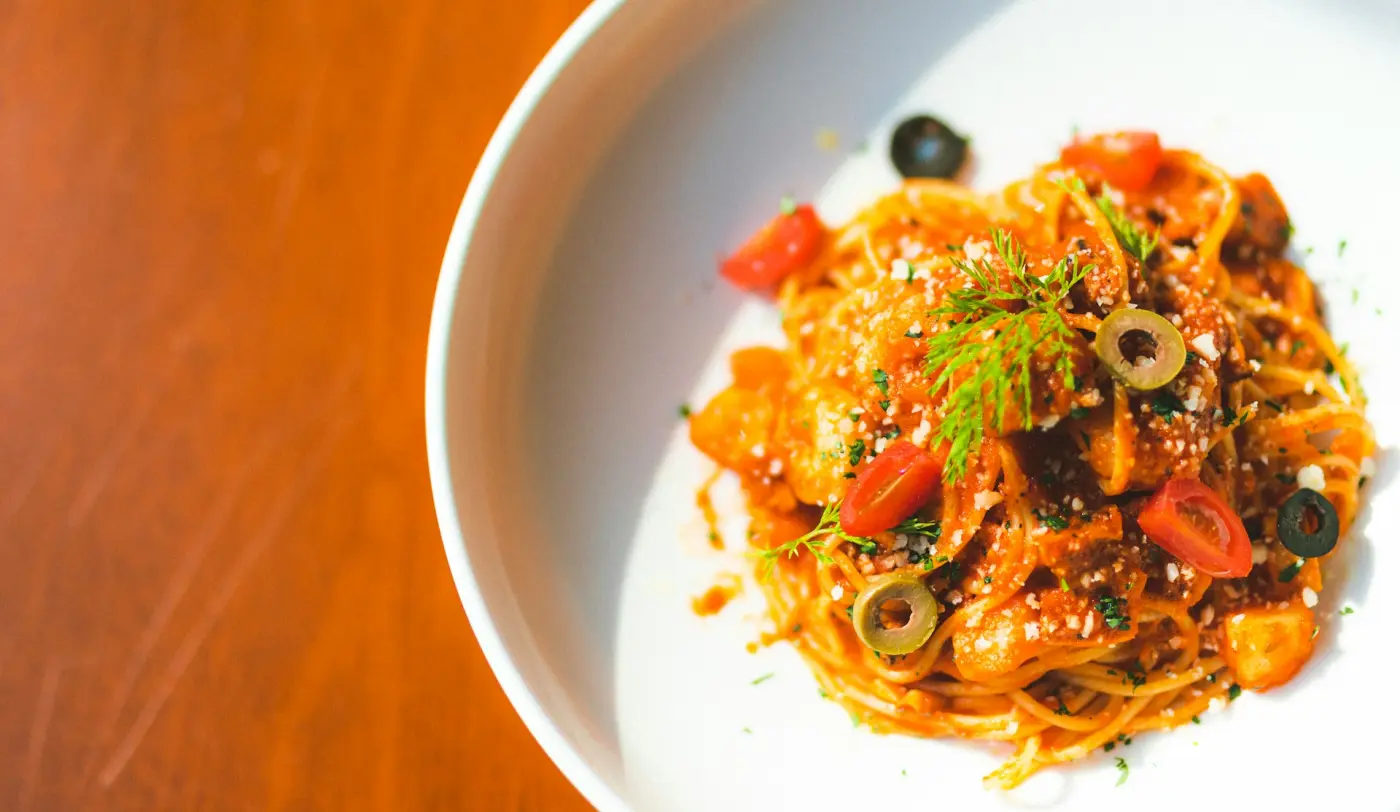 A bowl of spaghetti with tomato, olives and herbs on a wooden table